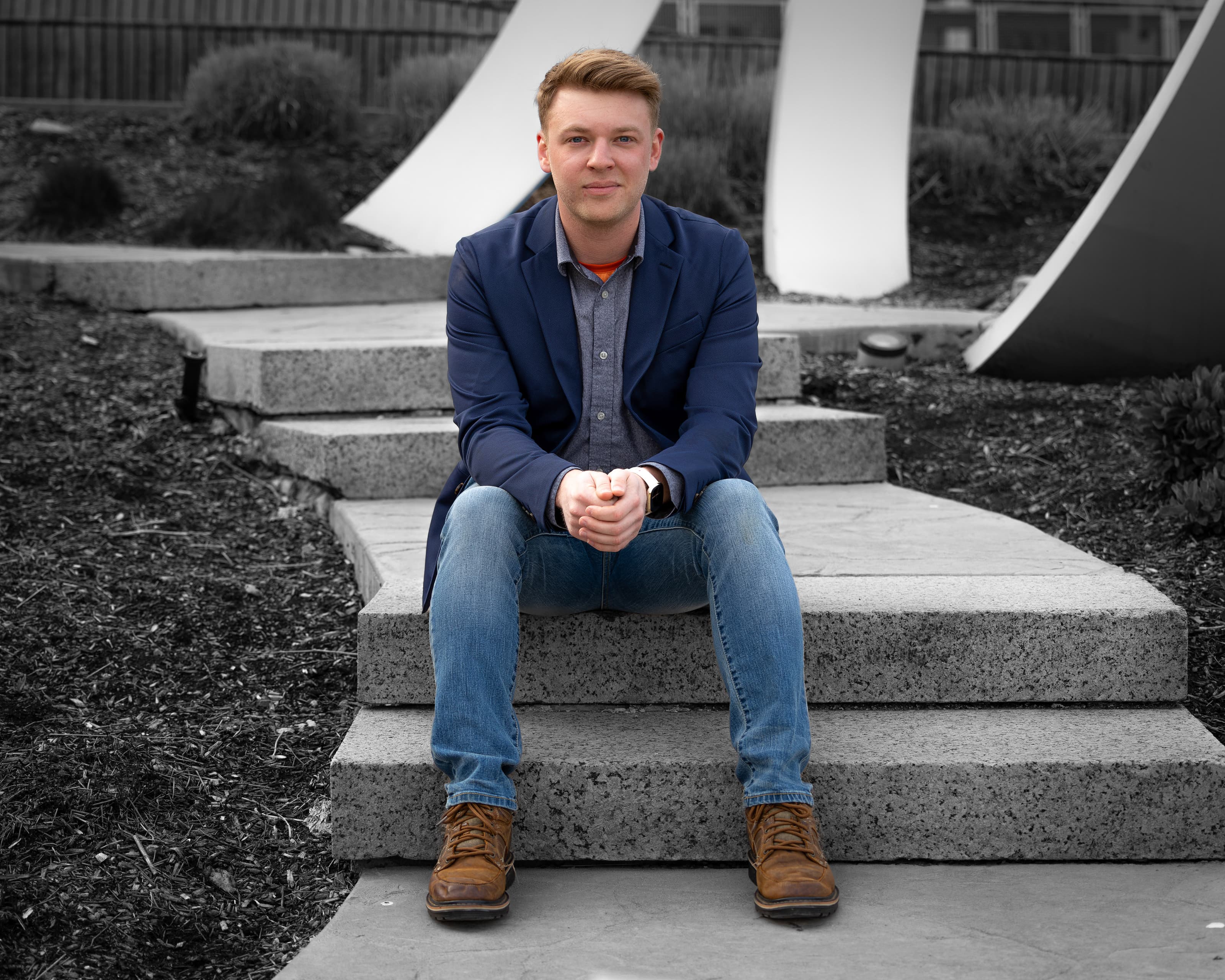 Brendan Goforth seated on stone steps in a navy blazer and jeans, in front of white sculptures.